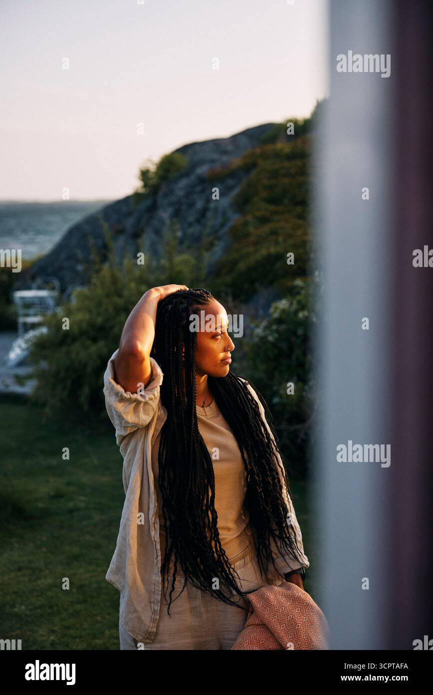 Besinnliche Frau mit Hand im Haar, die im Hinterhof des Ferienhauses steht, vom Fenster aus gesehen Stockfoto