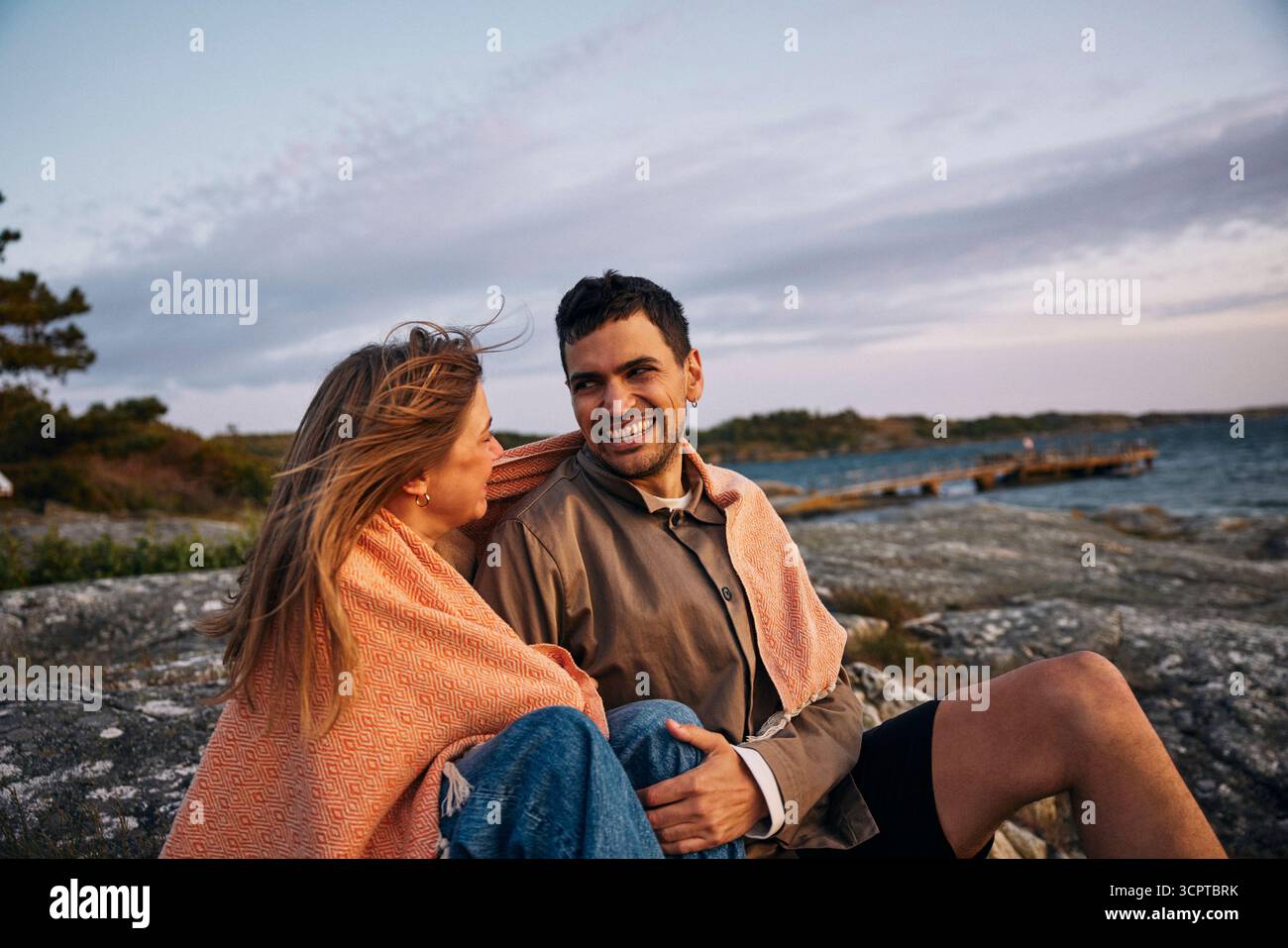 Lächelndes Paar, das in der Abenddämmerung auf einem Felsen sitzt, in einer Decke in der Nähe des Meeres Stockfoto