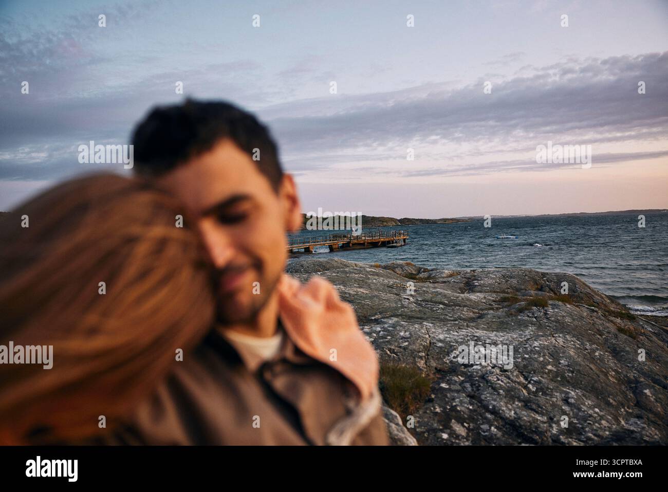 Defokussiertes, liebevolles Paar mit ruhiger Szene des Meeres in der Nähe von Felsen im Hintergrund in der Abenddämmerung Stockfoto