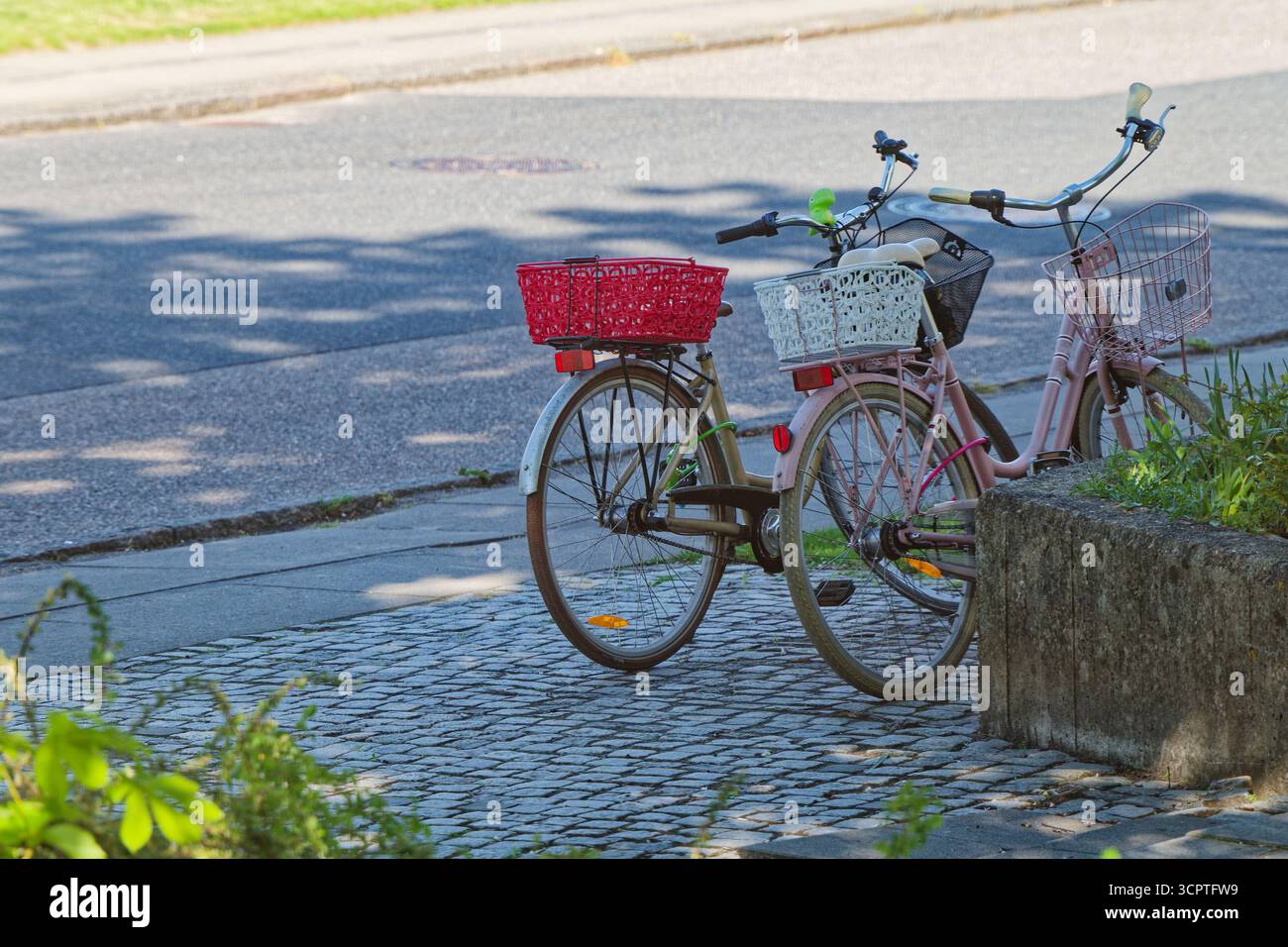 Zwei Fahrräder im Vintage-Stil, eines rosa und eines beige, ruhen zusammen auf dem Bürgersteig, jedes mit Korb vorne und hinten und cremefarbenen Reifen, die an das Gebäude erinnern Stockfoto