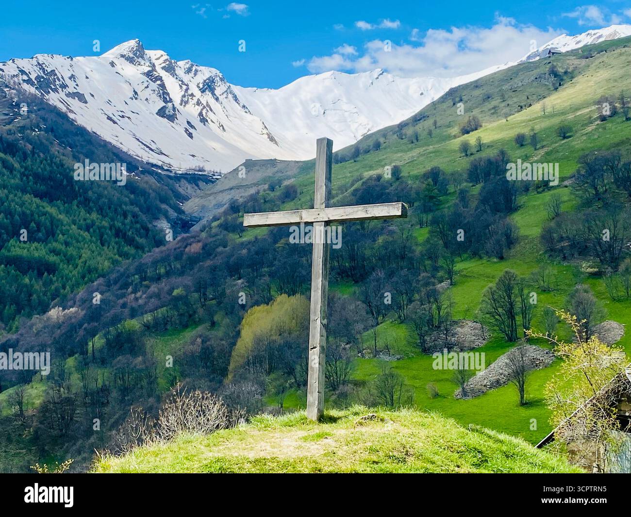 Holzkreuz auf Grassy Hill mit schneebedeckten Bergen und blauem Himmel in friedlicher Landschaft - Smartphone-aufgenommenes Stockfoto