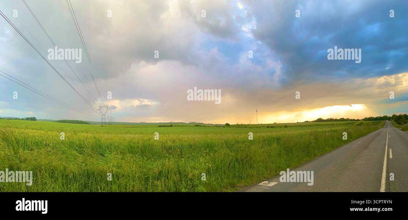 Gerader Landweg mit Stromleitungen und Sonnenuntergang, der durch die Sturmwolken über Green Fields führt - Smartphone-aufgenommenes Stockfoto