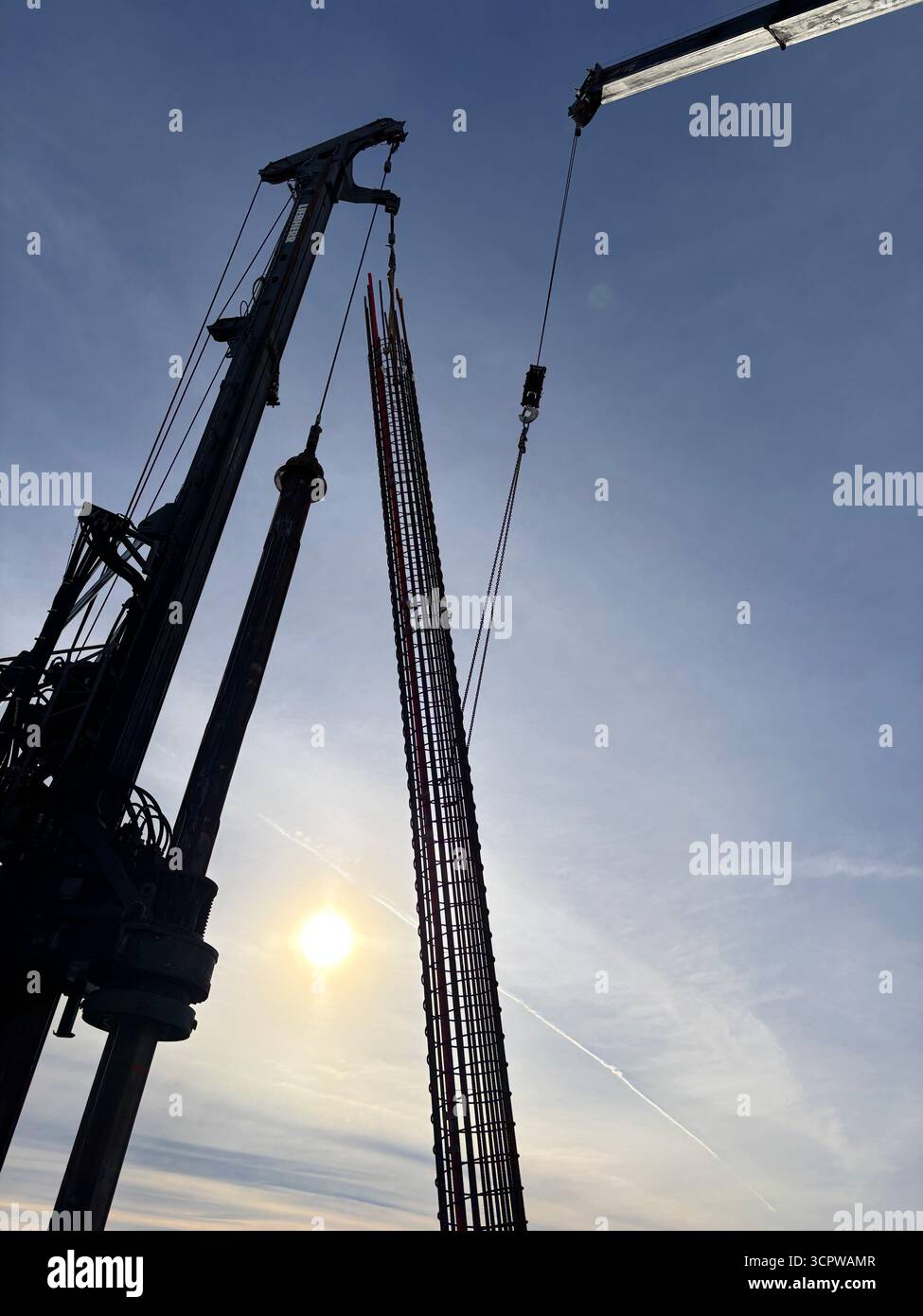 Kran hebt Bewehrungsstäbe an einem sonnendurchfluteten Himmel an und zeigt Bauarbeiten mit modernen Maschinen und klarem Horizont. - Smartphone-aufgenommenes Stockfoto