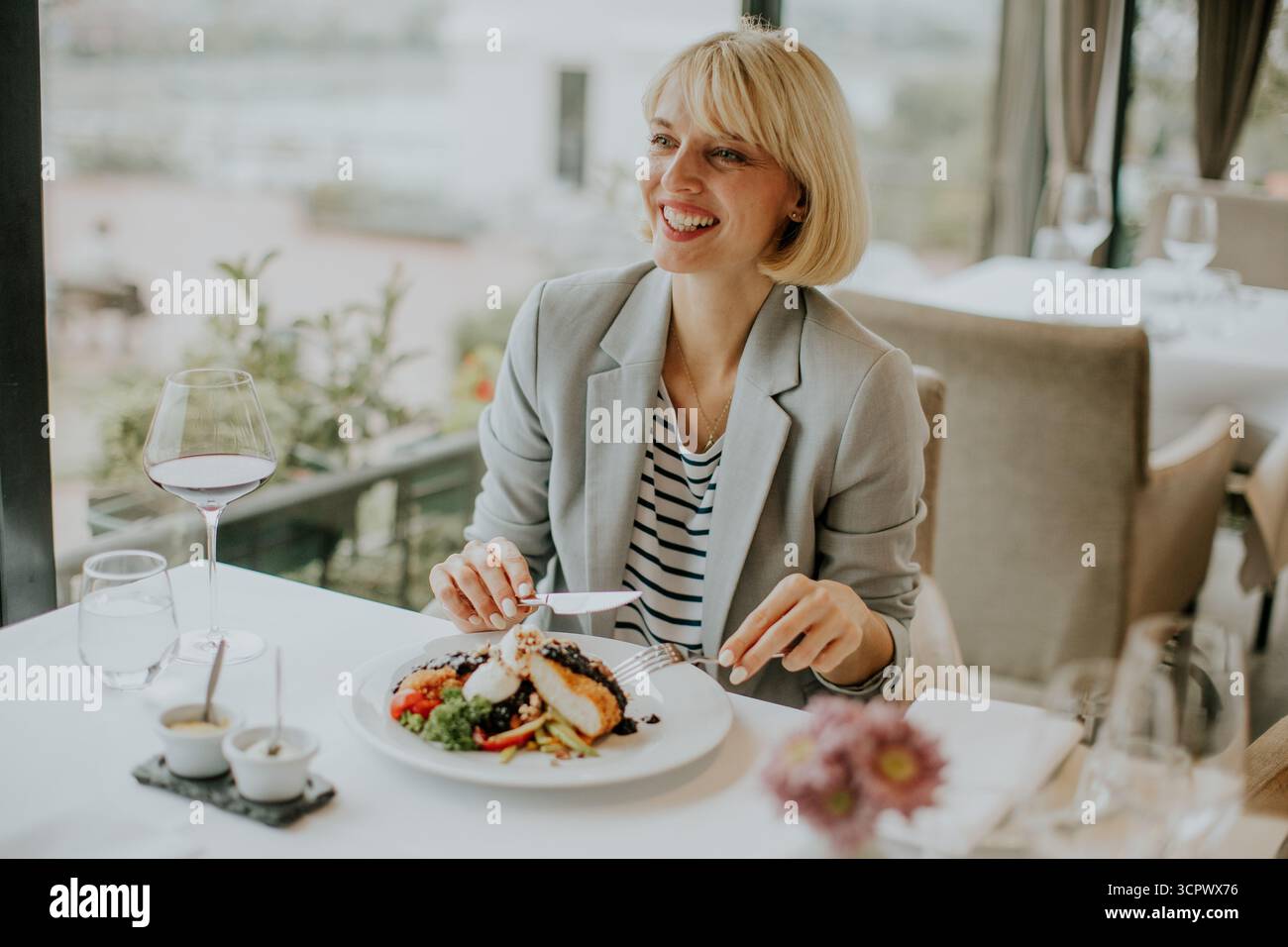 Eine lächelnde Frau sitzt an einem Tisch in einem schicken Restaurant und genießt ein wunderschön präsentiertes Gericht. Die Einstellung verfügt über große Fenster, die eine helle Sonnenliege ermöglichen Stockfoto