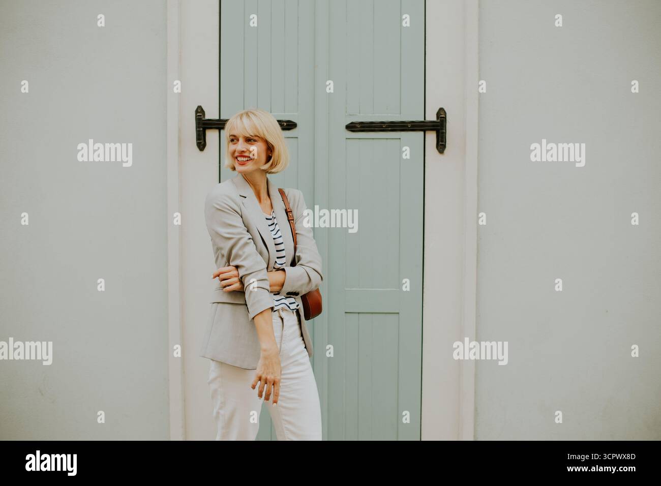 Eine modische Frau steht stolz vor einer hellblauen Tür. Ihr Outfit besteht aus einem Blazer und einem gestreiften Hemd, das fröhliche und selbstbewusste S einfängt Stockfoto
