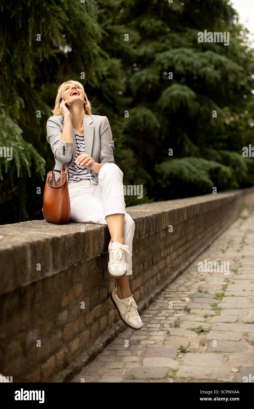Eine Frau mit Blazer und lässigem Outfit sitzt auf einer Steinmauer in einem grünen Park. Sie lächelt, während sie am Telefon spricht, und genießt einen fröhlichen Moment Stockfoto