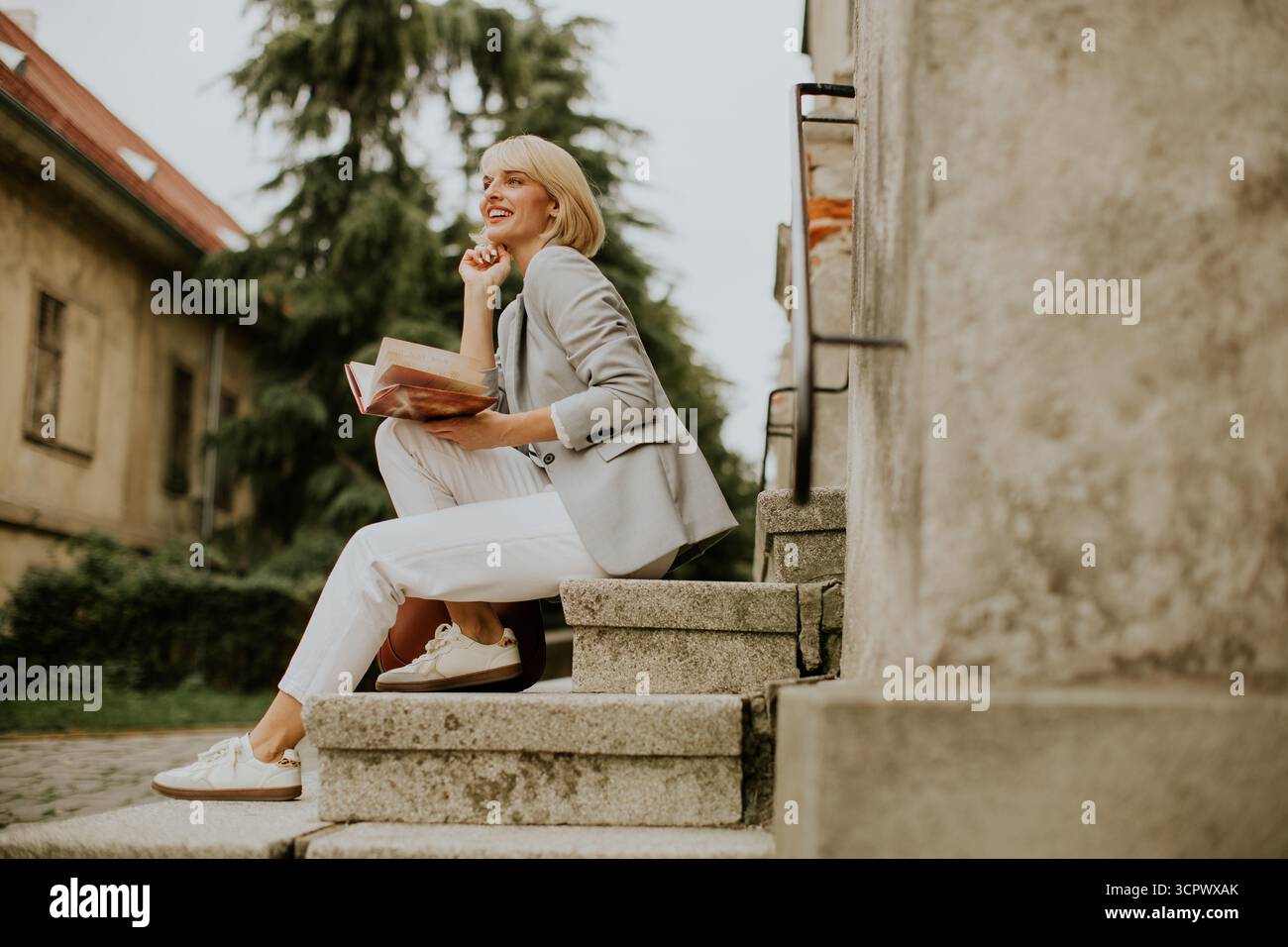 Eine Frau sitzt auf Steintreppen in einem ruhigen Innenhof und genießt ein Buch in legeren Kleidern. Üppig grüne Bäume bieten eine beruhigende Kulisse in der Th Stockfoto