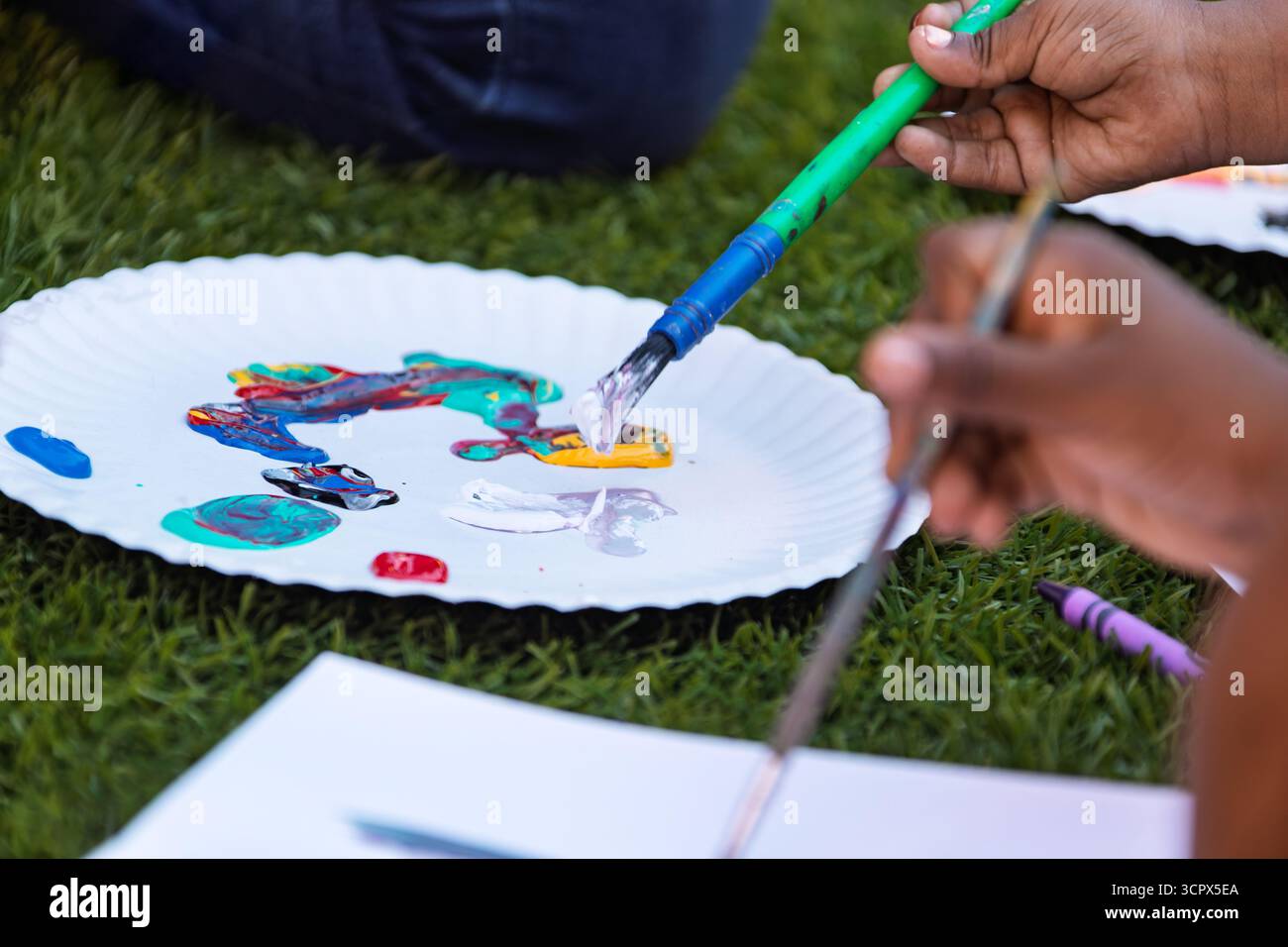 Hände afrikanischer Kinder, die Farben mit einem Pinsel auf einem Teller mischen, auf dem Gras draußen Stockfoto
