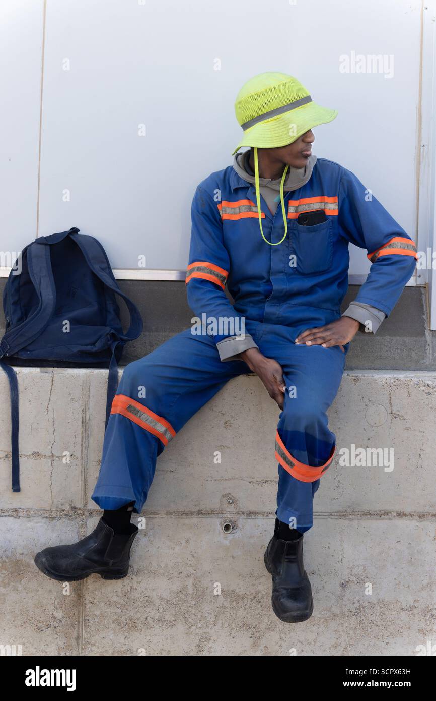 Einzelner afrikanischer Bauarbeiter, der draußen auf dem Hof eines Lagers in Arbeitskleidung mit Hut und Rucksack vor einem Lager stand Stockfoto