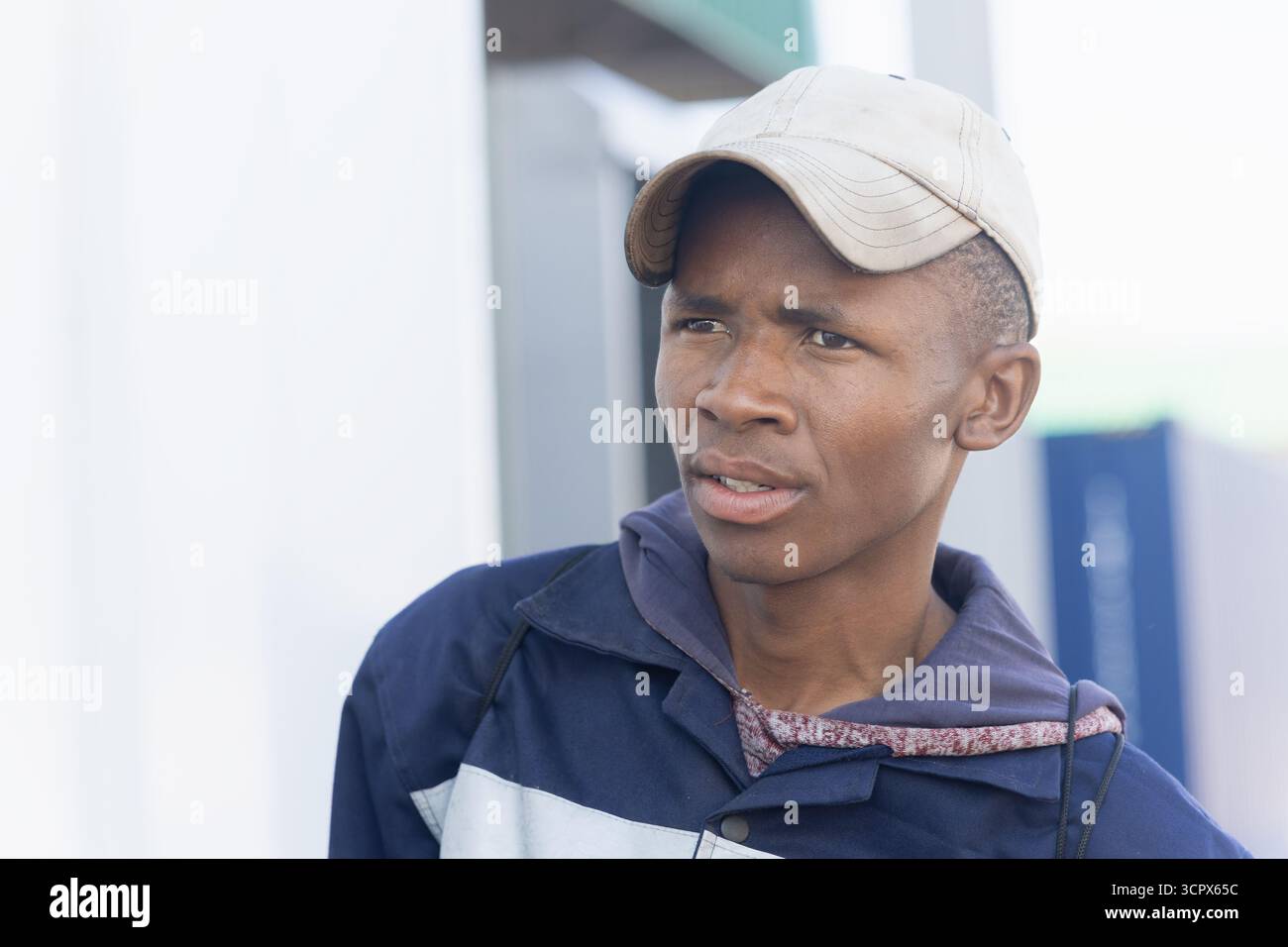 Einzelner afrikanischer Arbeiter, der draußen auf dem Hof eines Lagerhauses neben der Gebäudewand läuft und einen Hut trägt Stockfoto