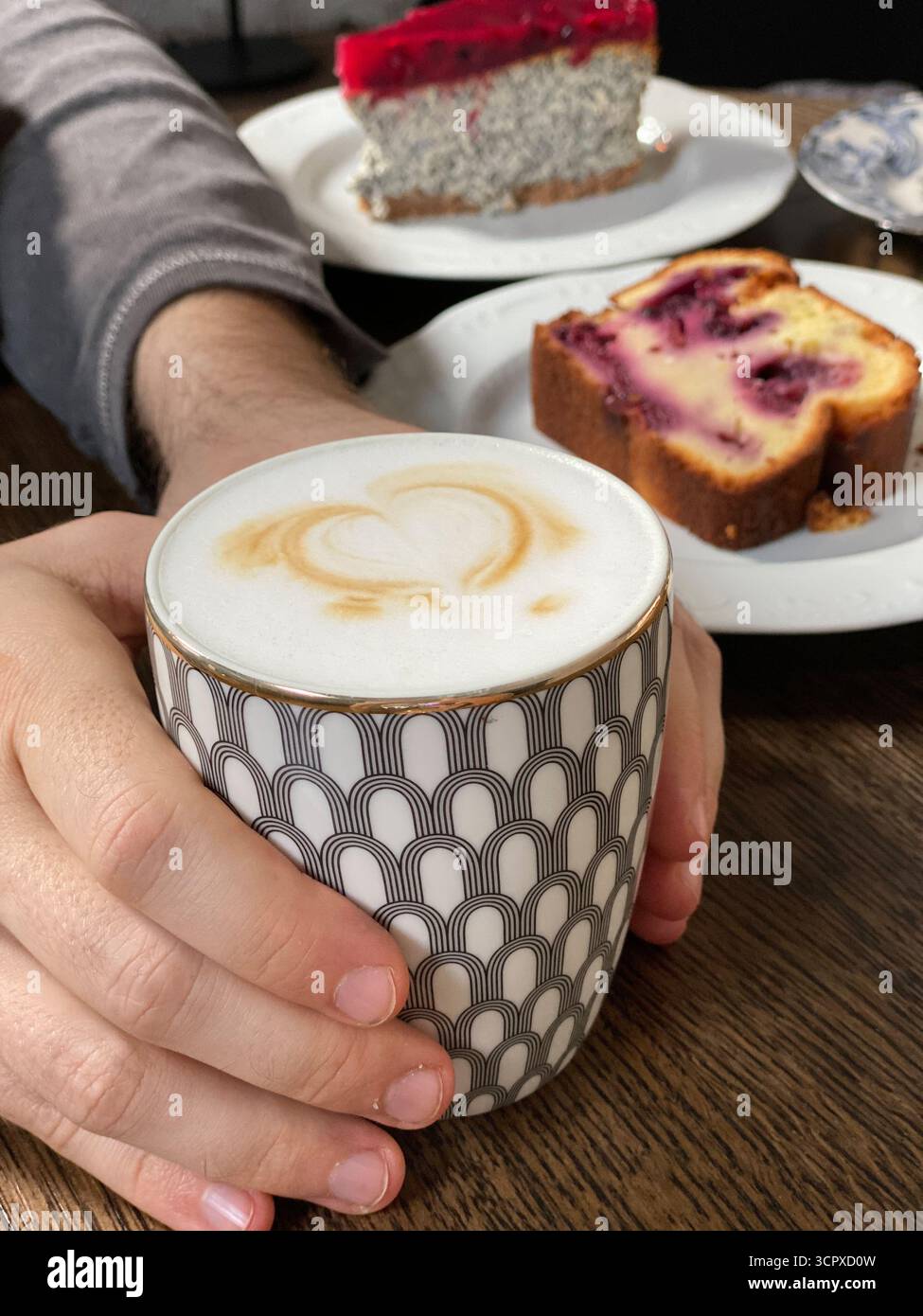 Hände halten Kaffeetasse mit Herzschaum auf Tisch mit Beerenkuchen und Mohnkuchen. Mobiles Foto Stockfoto