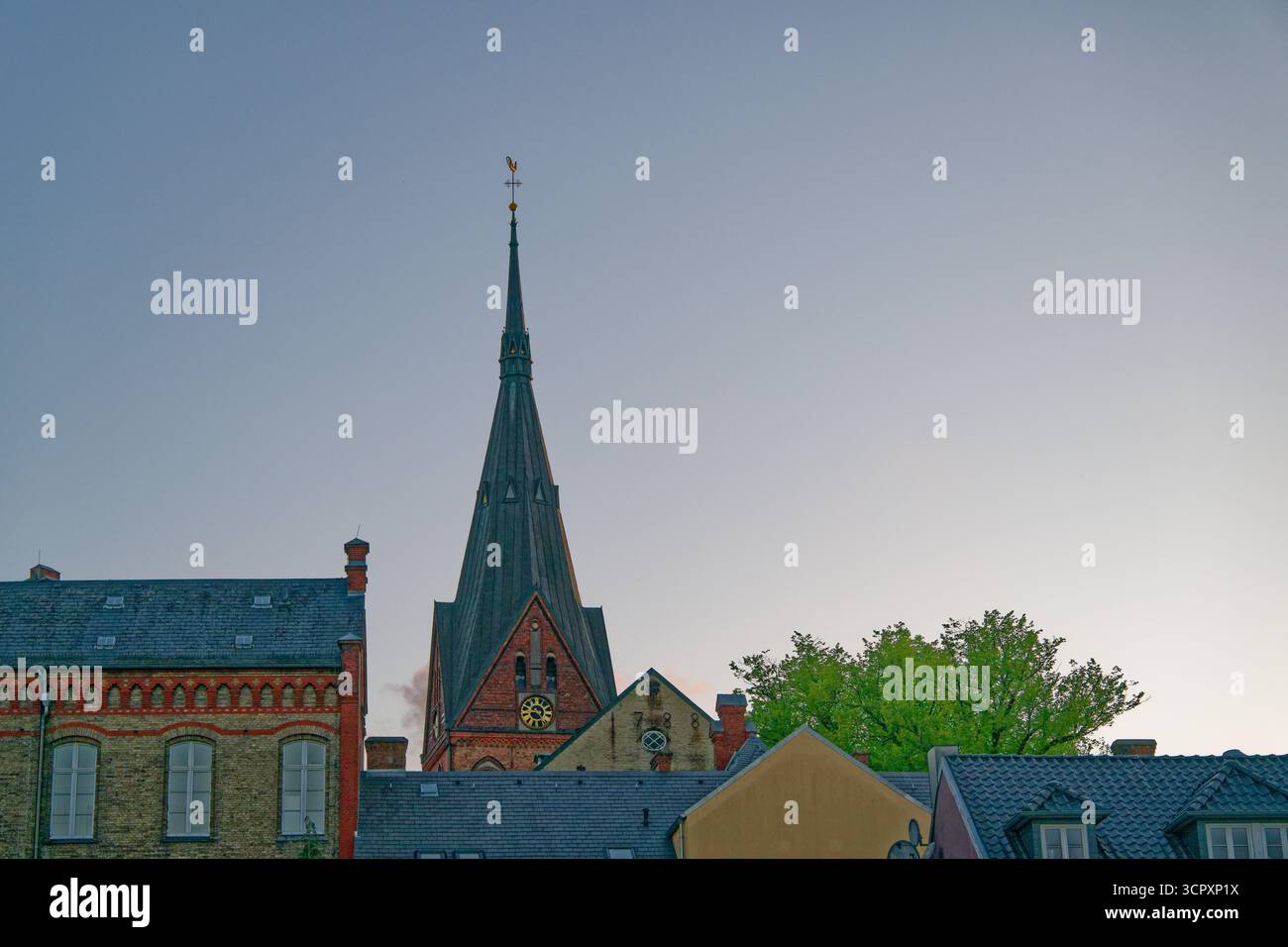 Der Flensburger Kirchturm erhebt sich mit einem dunklen Dach und einem Metallhahn auf der Wetterfahne, die den Wind markiert. Rote Backsteinmauern umrahmen einen großen goldenen Cloc Stockfoto