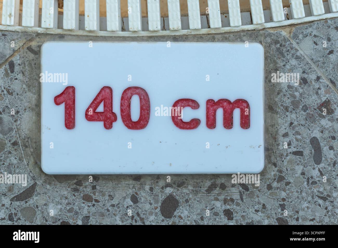 Nahaufnahme eines weißen Schildes mit rotem Text „140 cm“, das auf einem Steinboden in der Nähe eines weißen Zauns platziert wurde. Stockfoto