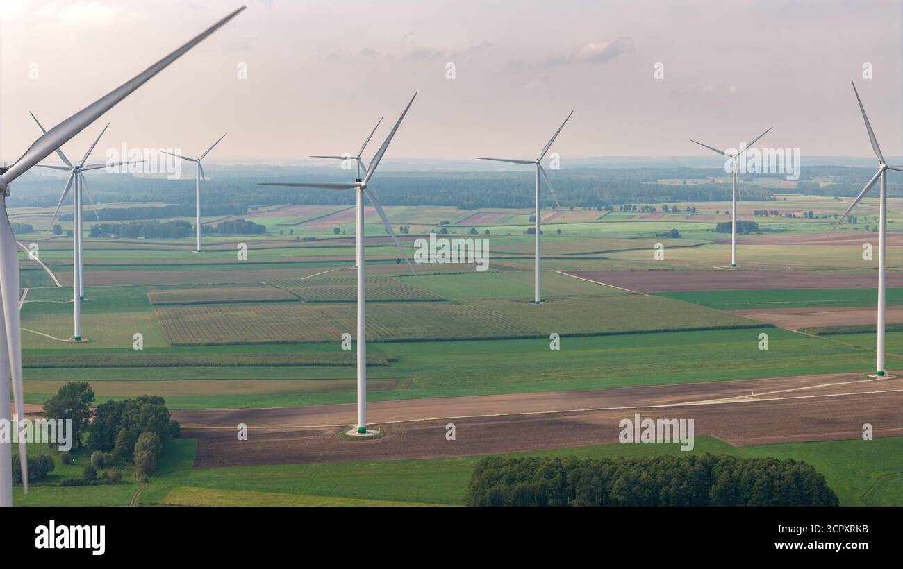 Große Windkraftanlagen stehen auf grünem und braunem Ackerland, mit Baumgruppen und einem trüben Horizont unter freiem Himmel. Stockfoto