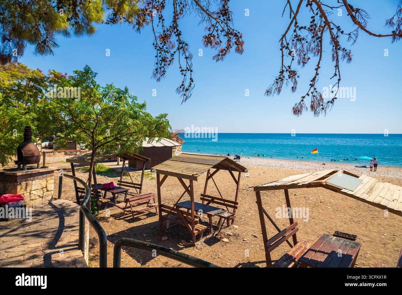 Strandszene mit Picknicktischen unter schattigen Strukturen, blauem Meer, klarem Himmel und einigen Leuten in der Ferne. Stockfoto