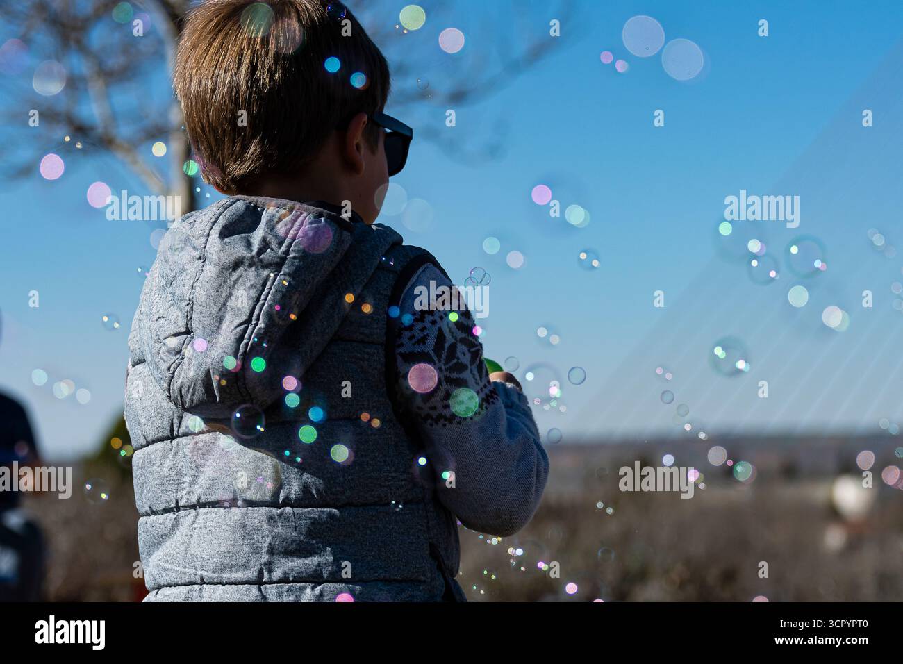 Ein kleines Kind mit Sonnenbrille spielt an einem hellen, klaren Tag in einem Park mit bunten Blasen. Stockfoto