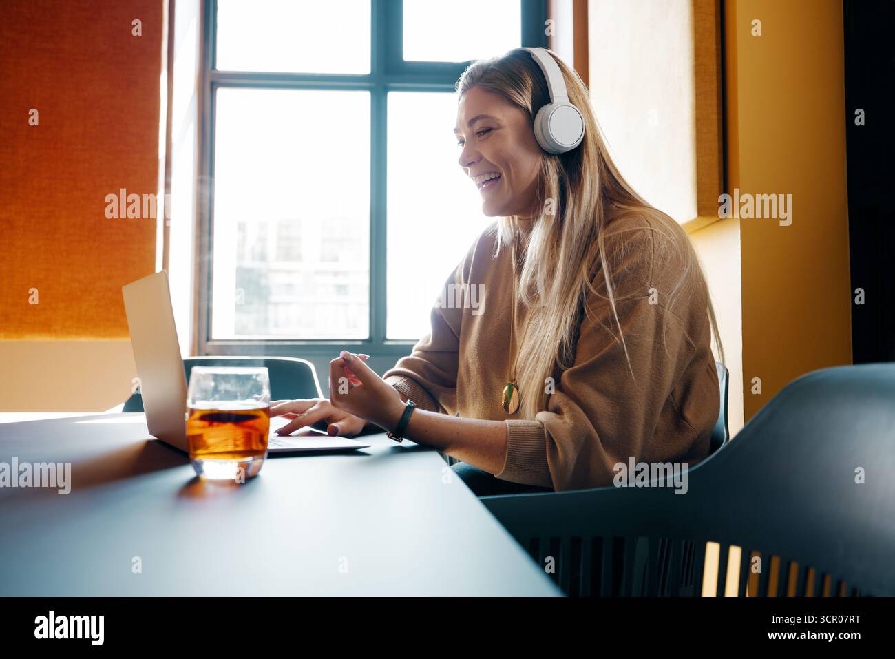 Eine junge Frau mit langen blonden Haaren sitzt drinnen und benutzt ihren Laptop, während sie Kopfhörer trägt. Sie lächelt und ist an einem Online-Meeting beteiligt Stockfoto