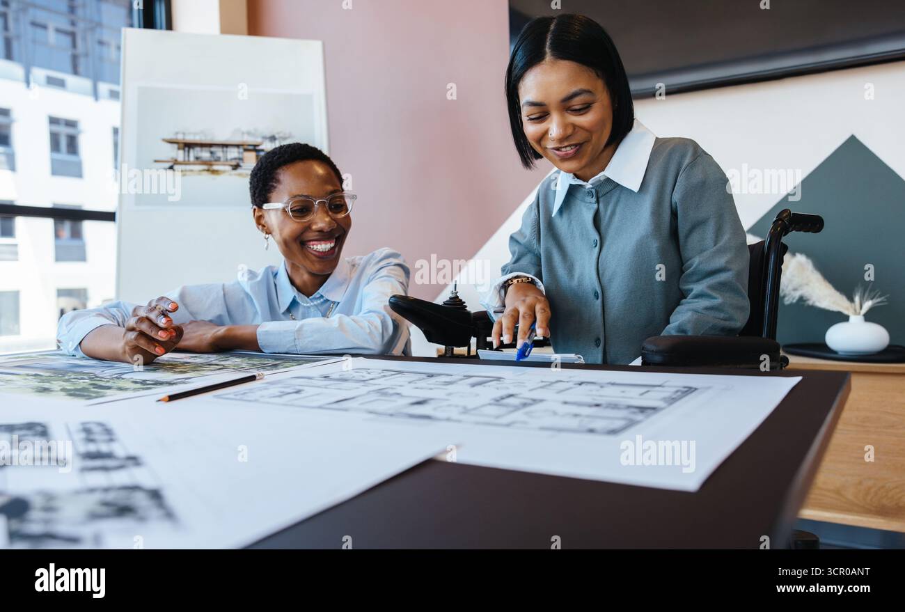 Zwei junge Frauen, eine davon im Rollstuhl, arbeiten an architektonischen Entwürfen und Entwürfen in einem Büro zusammen, das Kreativität und Vielfalt zum Ausdruck bringt. Stockfoto