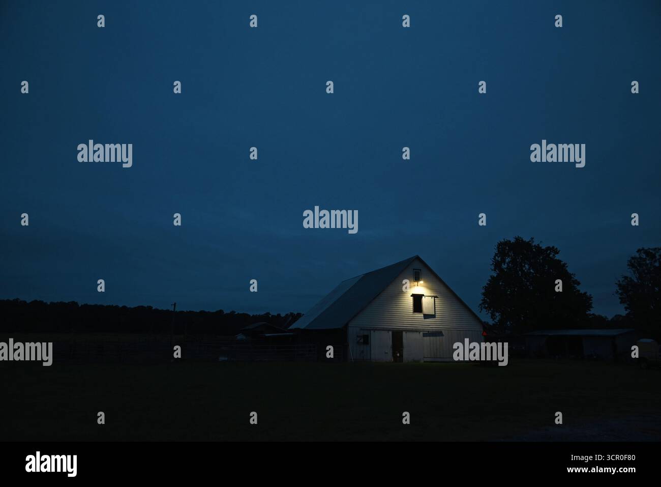 Eine Jahrhunderte alte weiße Scheune wird im Dunkeln von einem einsamen Licht vor Sonnenaufgang beleuchtet. Stockfoto