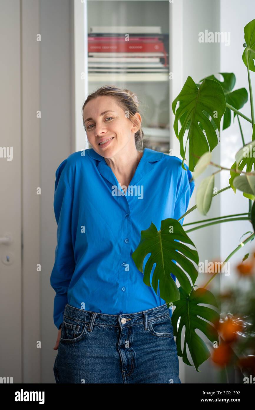 Porträt einer charmanten lächelnden Frau mittleren Alters steht neben Monstera-Blättern in einem gemütlichen Zuhause Stockfoto