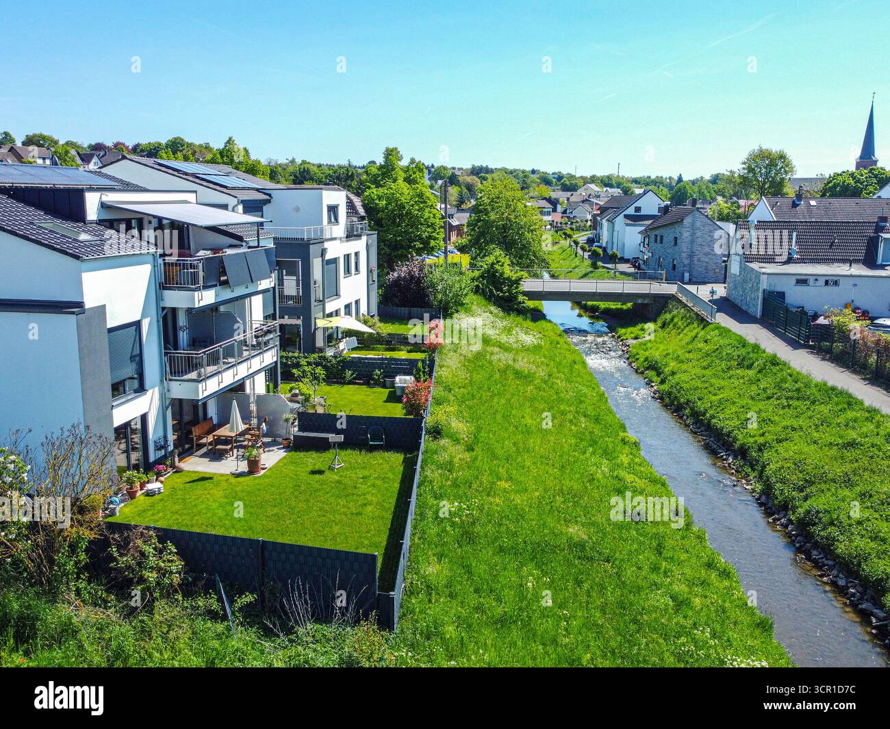 Moderne Häuser säumen ein ruhiges Flussbett mit gepflegten Rasenflächen. Helles Sonnenlicht beleuchtet die Szene und bietet eine friedliche Atmosphäre mit Grün Stockfoto