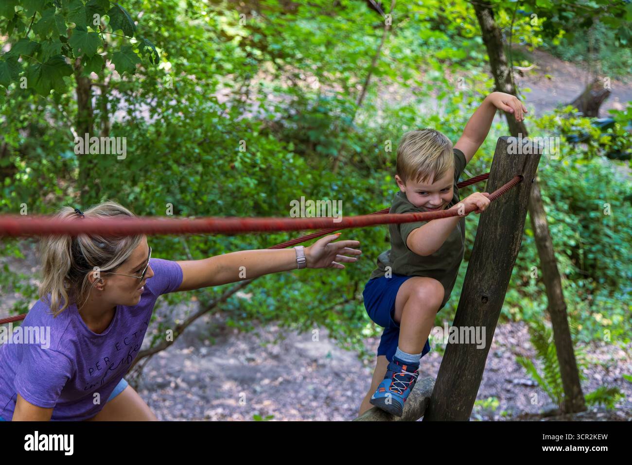 Kinder spielen auf Rutsche und Kletterseilen auf einem von Bäumen umgebenen Naturspielplatz. Stockfoto