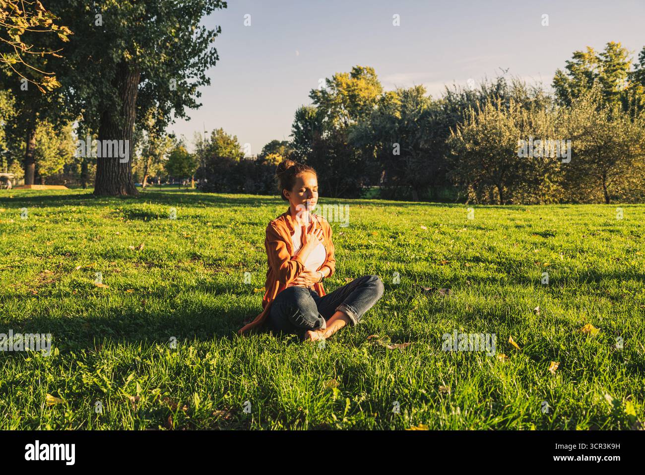Erwachsene Frau in lässigem Outfit, die nach der Arbeit in Lotuspose auf dem Gras im Stadtpark meditiert. Stockfoto