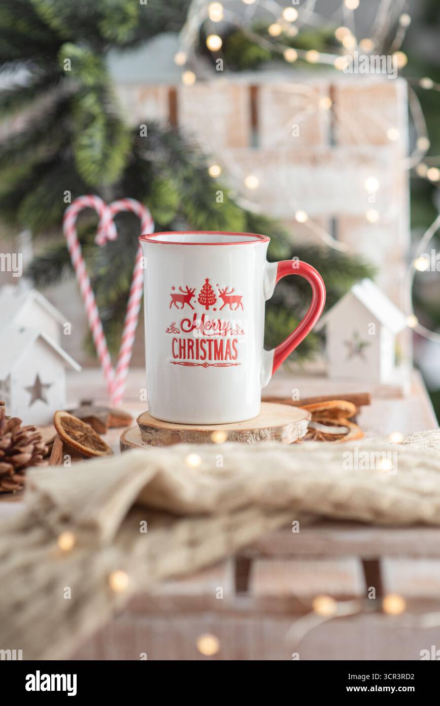 Keramikbecher im Weihnachtsdesign auf Holzuntersetzer, umgeben von festlichen Elementen wie Tannenzapfen und Lichtern, die einen warmen Weihnachtsgeist wecken Stockfoto