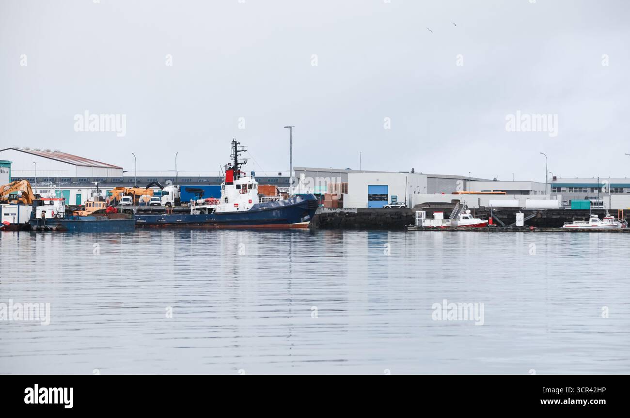 Der Industriehafen an einem Kai zeigt ein großes blaues Schiff, Kräne, Lagerhäuser und kleine Boote. Reykjavik, Island Stockfoto