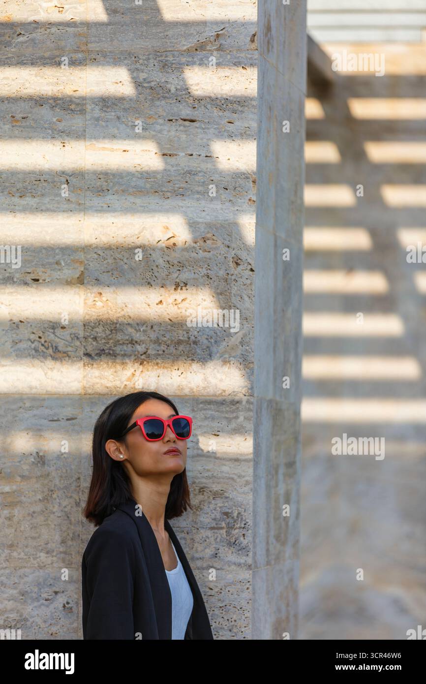 Frau in Sonnenbrille steht vor einer sonnendurchfluteten Steinmauer mit Schatten. Spanien Stockfoto