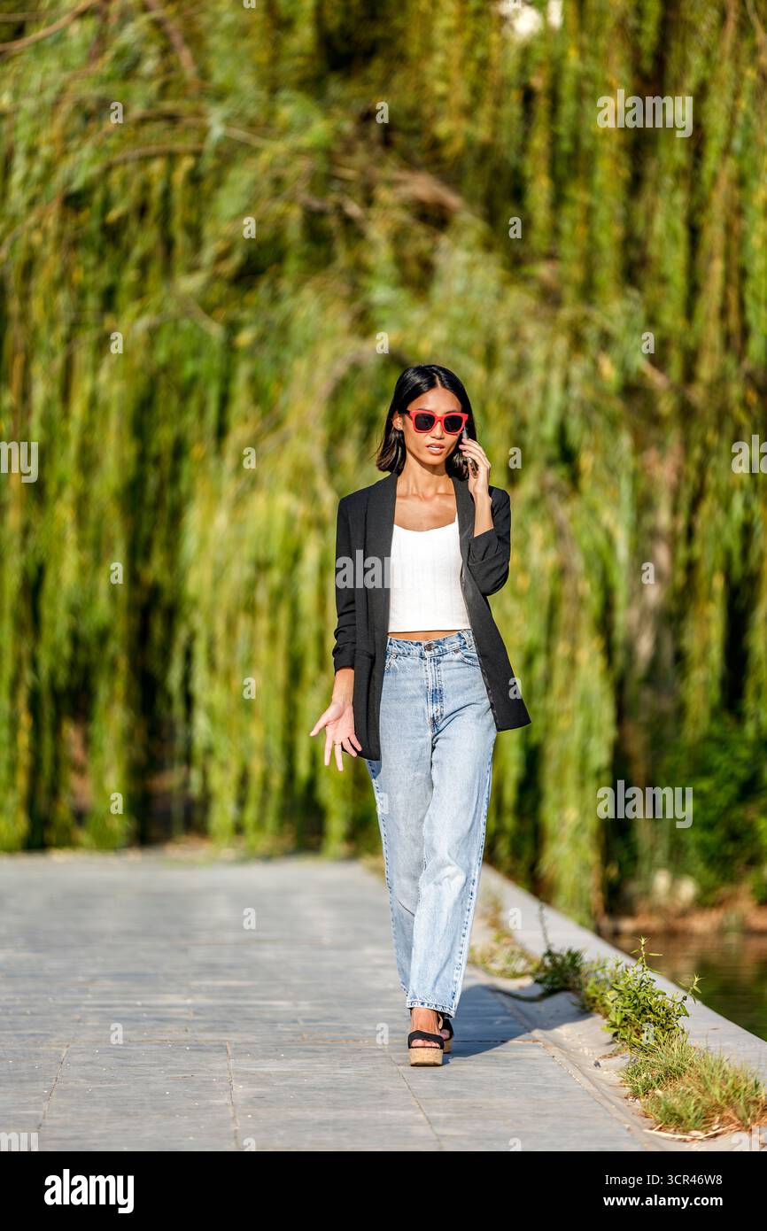 Frau in lässiger Kleidung, die draußen läuft, rote Sonnenbrille trägt und ein Smartphone benutzt. Spanien Stockfoto