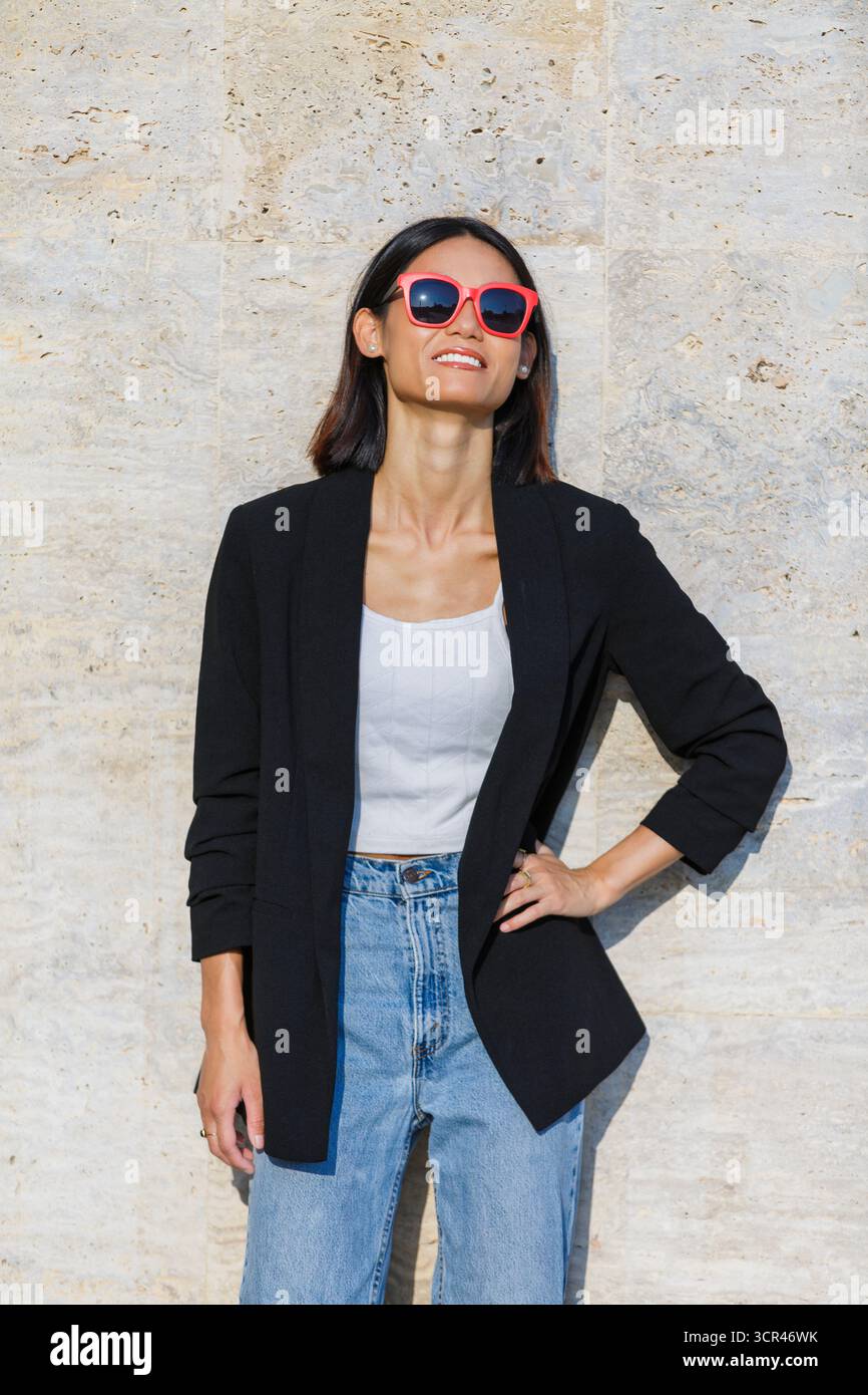 Frau in Sonnenbrille und lässigem Outfit lächelt, während sie an einer Steinmauer steht. Spanien Stockfoto