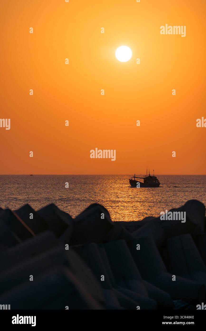 Silhouette eines Bootes, das bei Sonnenuntergang auf dem Meer segelt, mit orangefarbenem Himmel und Betonblöcken. Phu Quy, Vietnam Stockfoto