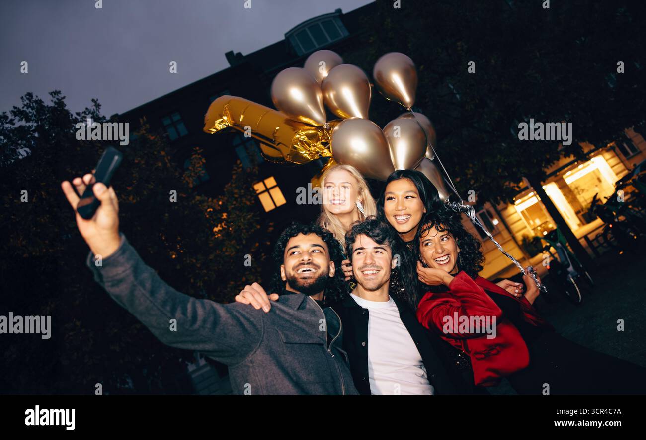 Eine fröhliche Gruppe von Freunden fängt den Moment mit einem Selfie ein und feiert gemeinsam. Sie halten Ballons in einer städtischen Umgebung am Abend, Crea Stockfoto