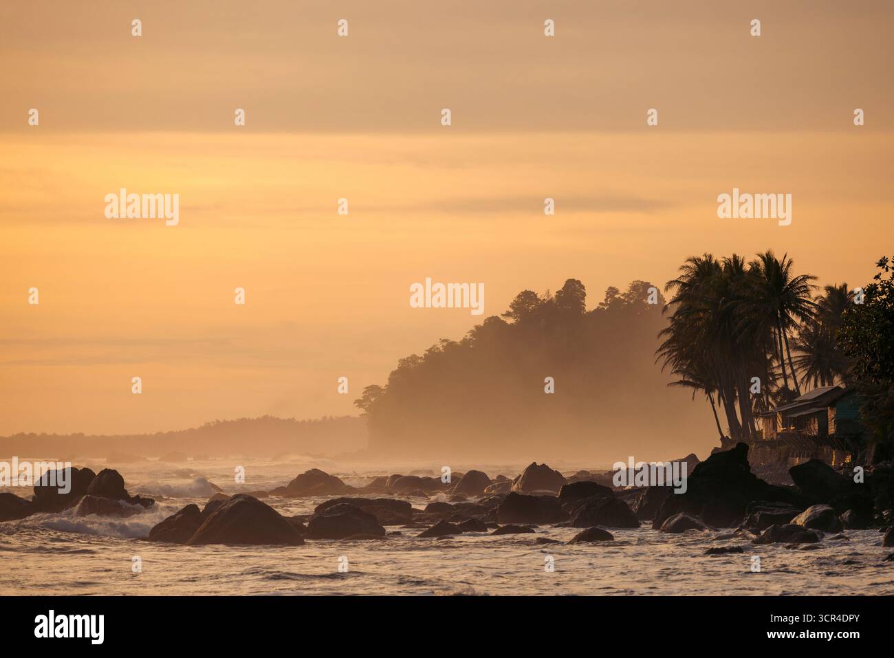 Sonnenuntergang über einem felsigen Strand mit Silhouetten Palmen und ruhigen Wellen in der Ferne. Krui, Lampung, Sumatra, Indonesien Stockfoto