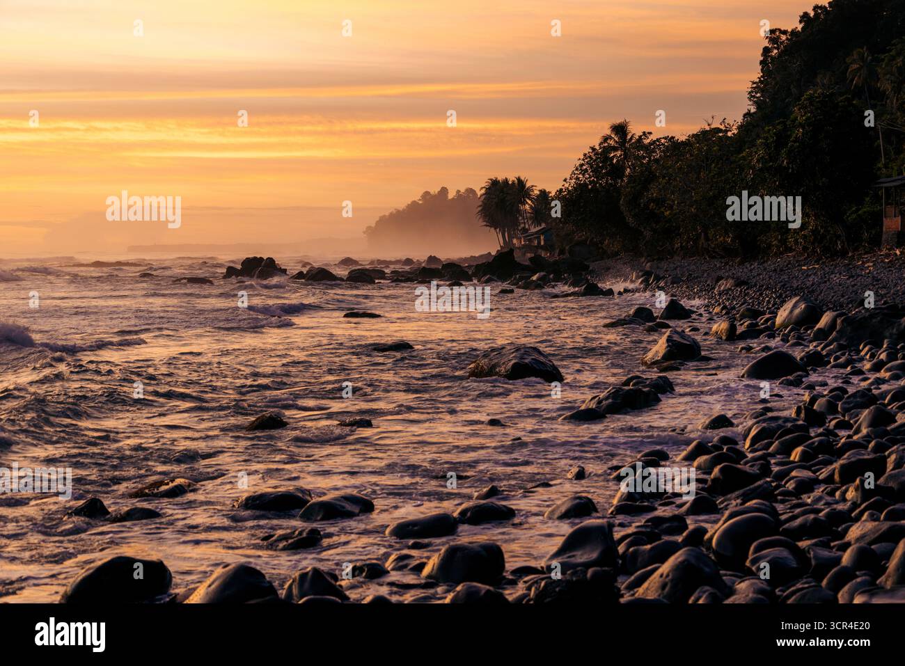 Dramatischer Sonnenuntergang an der Küste mit felsiger Küste und Silhouetten von Bäumen vor orangem Himmel. Krui, Lampung, Sumatra, Indonesien Stockfoto