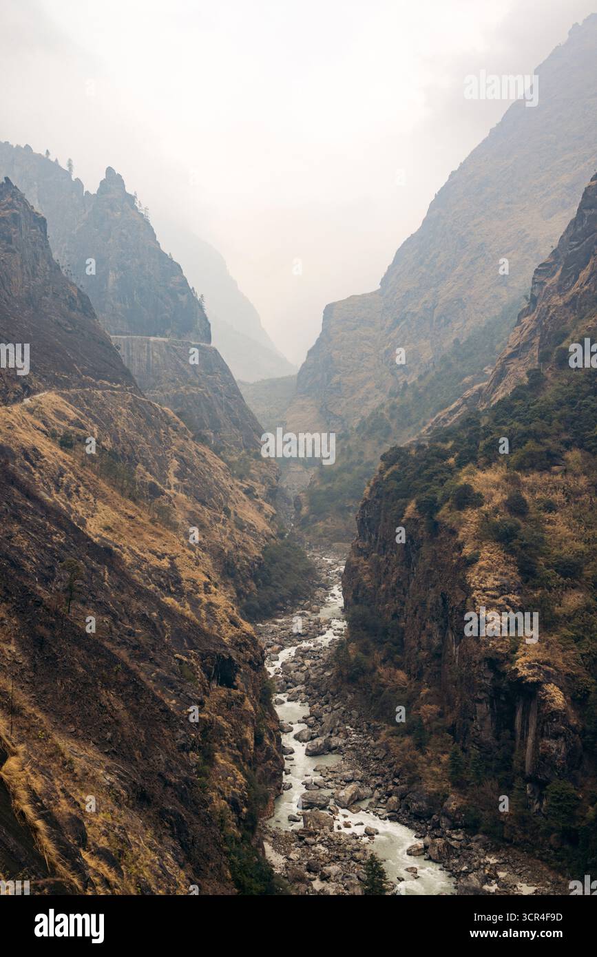 Majestätisches Bergtal mit einem sich windenden Fluss und steilen, zerklüfteten Klippen unter einem nebeligen Himmel. Straße F36 von Annapurna in der Provinz Gandaki, Nepal Stockfoto