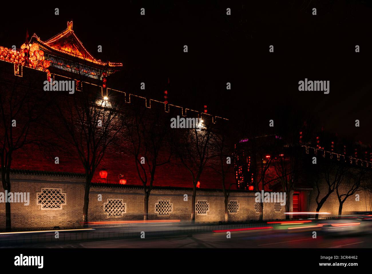 Historisches Gebäude mit beleuchteten roten Lichtern bei Nacht, Bäume werfen Schatten auf die Wände. Befestigungsanlagen von Xi'an, China Stockfoto