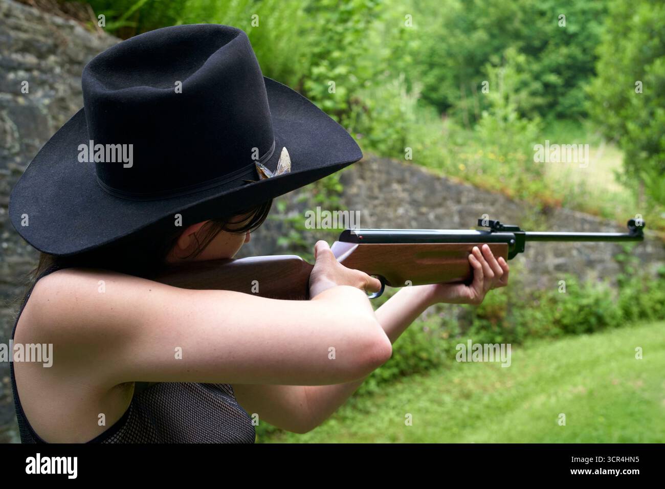 Frau mit schwarzem Hut, die draußen mit einem Gewehr auf eine von Grün umgebene Zielreichweite zielt. Stockfoto