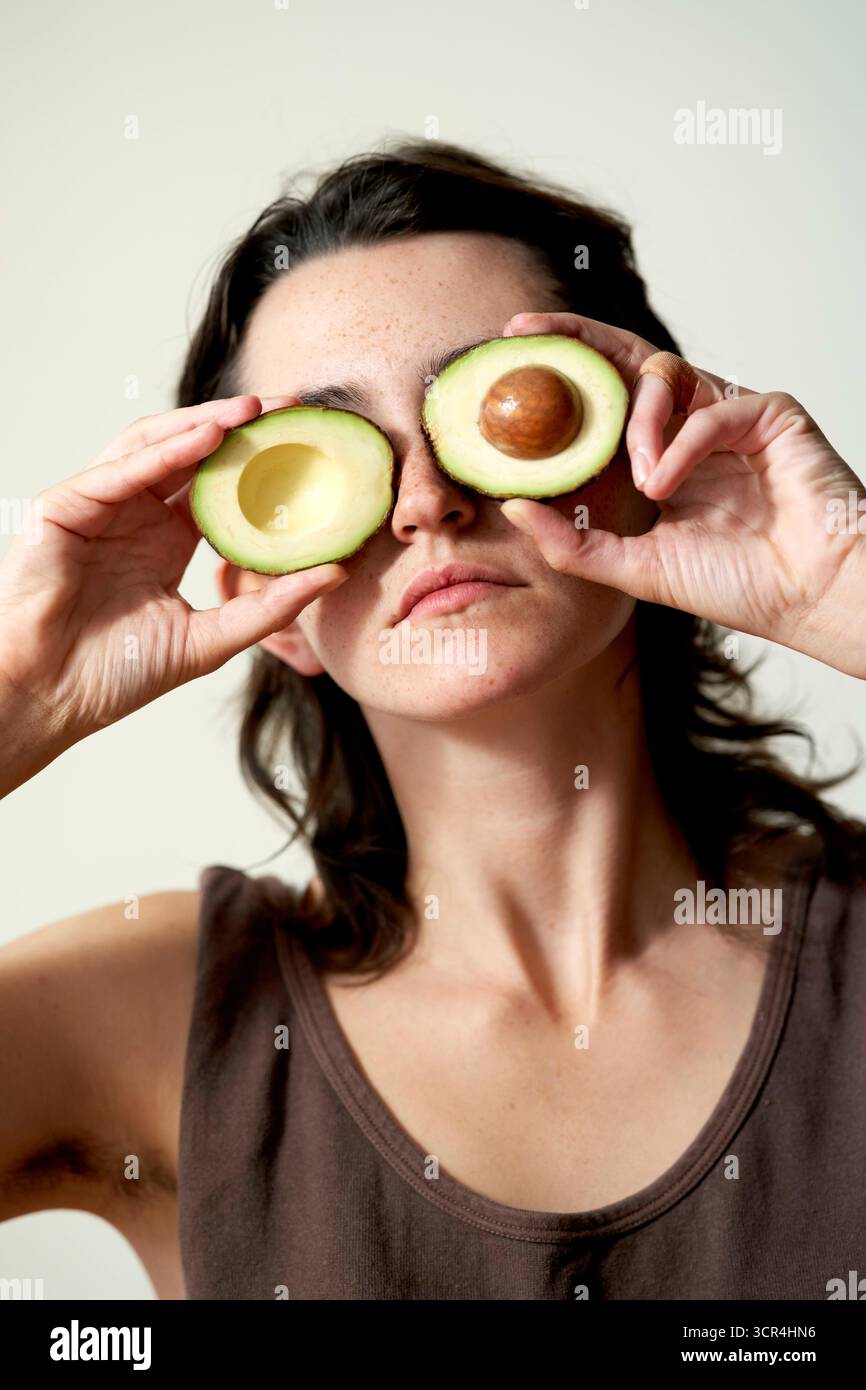 Frau, die Avocadohälften über Augen hält wie eine Brille vor neutralem Hintergrund. Stockfoto