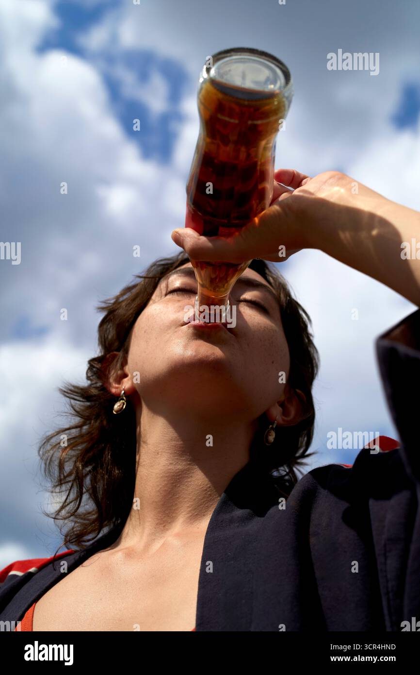 Frau trinkt aus einer Glasflasche vor einem bewölkten Himmel. Stockfoto