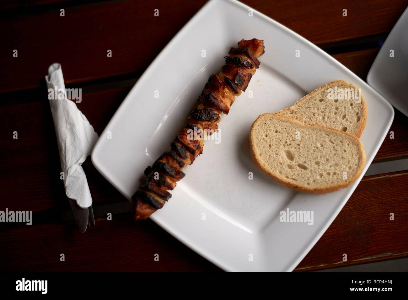 Gegrillter Kebab mit zwei Brotscheiben auf einem quadratischen weißen Teller, Serviette mit Besteck daneben. Stockfoto