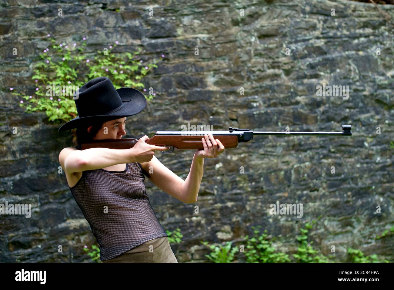 Frau mit schwarzem Hut, die draußen mit einem Gewehr auf eine von Grün umgebene Zielreichweite zielt. Stockfoto