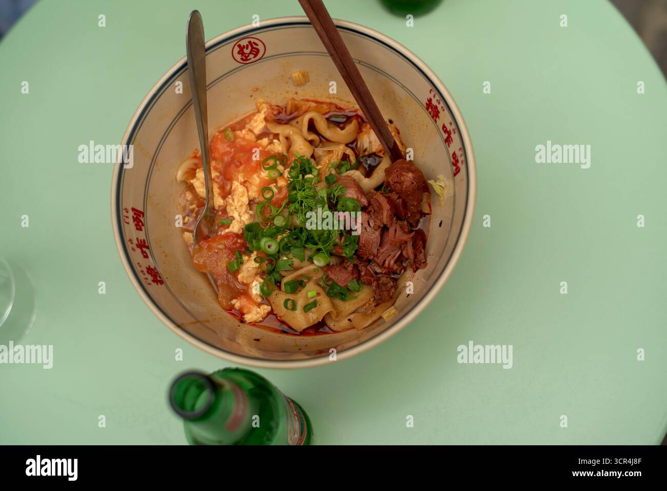 Scharfe Nudelsuppe in einer dekorativen Schüssel auf einem hellgrünen Tisch mit einer Flasche daneben. Brüssel, Belgien Stockfoto
