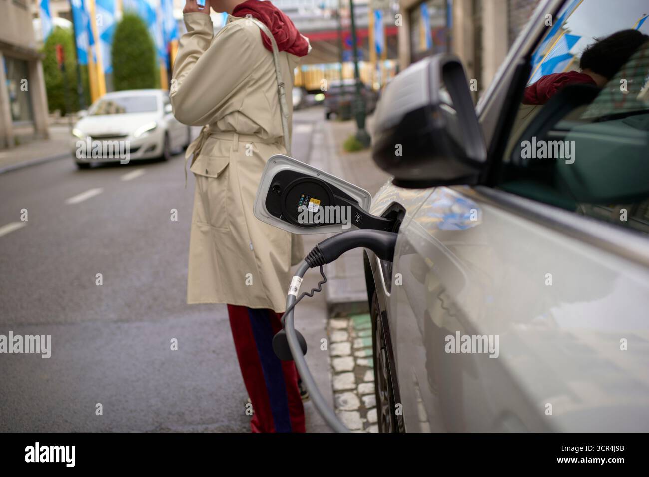 Elektroauto, das auf der Straße aufgeladen wird, während eine Person in legerer Kleidung ein Telefon hält. Brüssel, Belgien Stockfoto