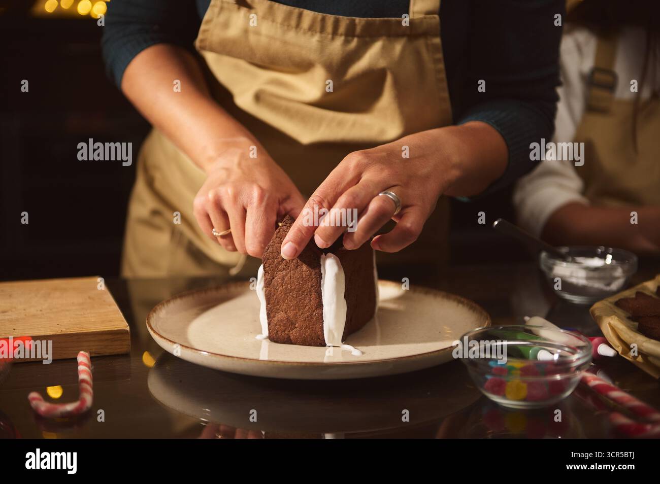 Ein Bäcker in einer Schürze ziert einen kleinen Kuchen mit weißer Glasur auf einem Teller, umgeben von Backwerkzeugen und farbenfrohen Dekorationen, die an warme Feiertagsküche erinnern Stockfoto