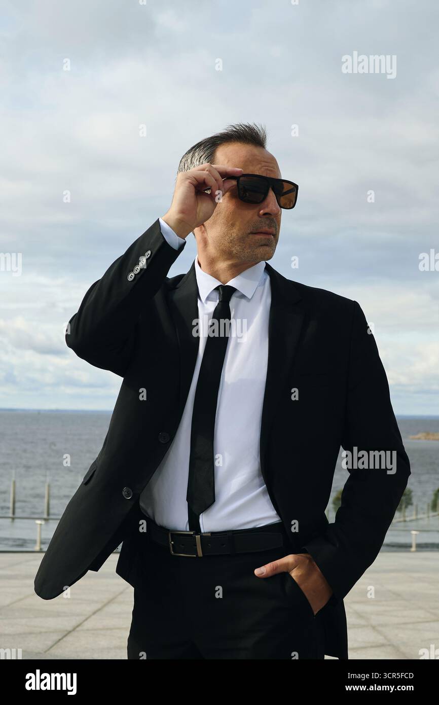 Kaukasischer Mann mittleren Alters, der Anzug und Sonnenbrille trägt, steht draußen in der Nähe des Ufers und passt die Sonnenbrille mit selbstbewusstem Ausdruck an, der von der Kamera wegblickt Stockfoto