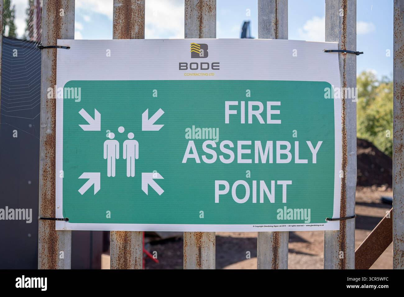Schließen Sie das grüne Schild „Fire Assembly Point“ von Bode Contracting Ltd., das an einem Stahlzaun angebracht ist und dahinter eine kommerzielle Baustelle in Großbritannien liegt. Stockfoto