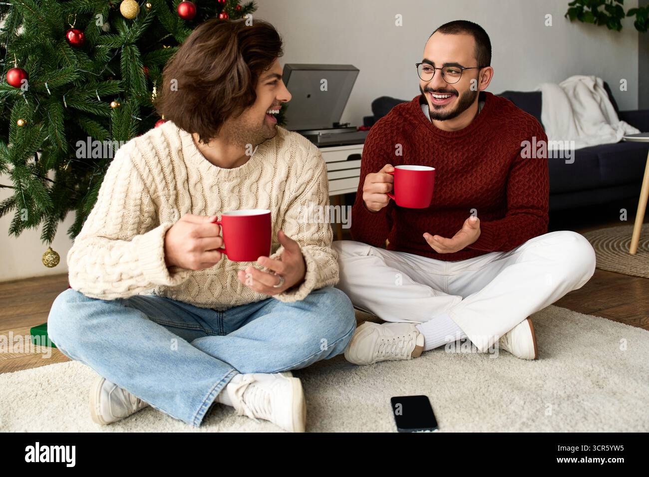 Zwei Männer teilen sich Lachen und Wärme, während sie zusammen auf einem gemütlichen Teppich sitzen und festliche Drinks genießen. Stockfoto