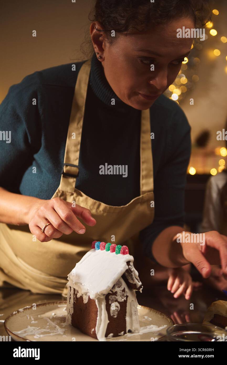 Eine Frau in einer beigefarbenen Schürze ziert ein Lebkuchenhaus und setzt Süßigkeiten in einem warmen, festlichen Zuhause. Die Urlaubsszene fängt Backkunst, Familienstimmung, Stockfoto